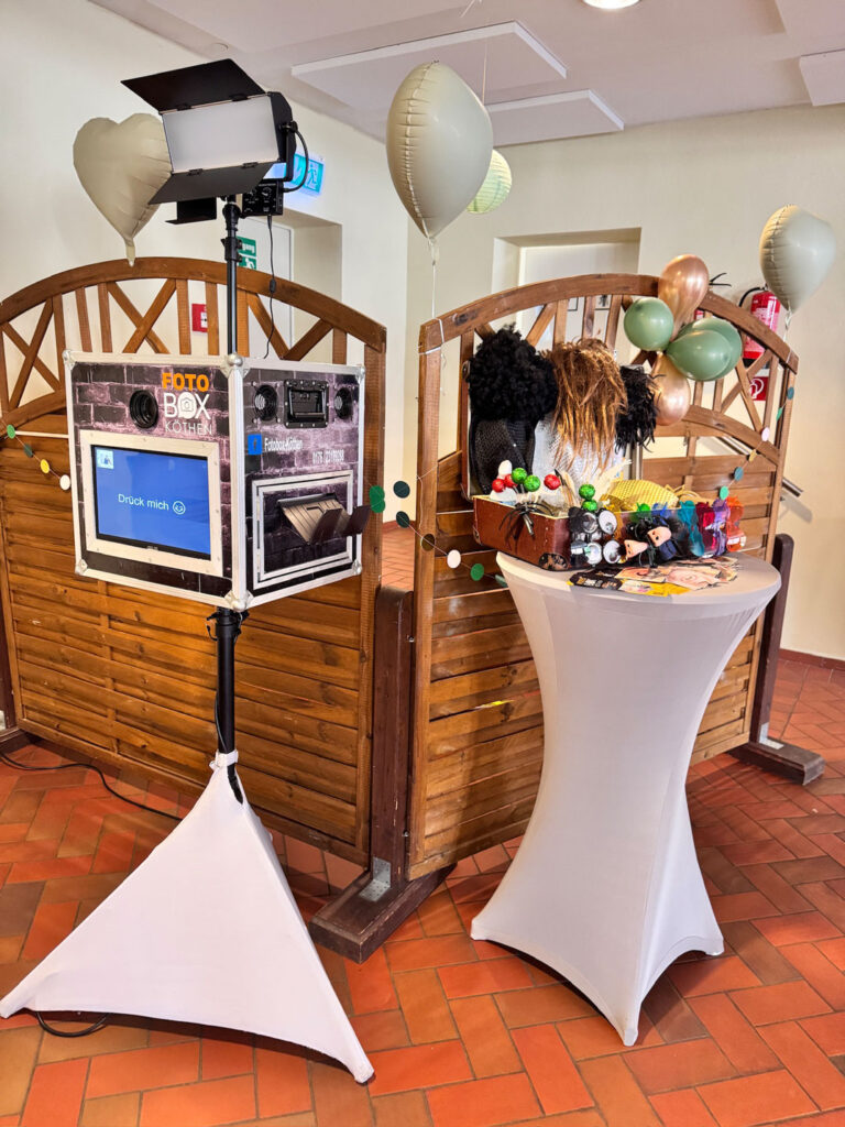 Fotobox mit Dekoration und Luftballons auf einer Feier, Stehtisch daneben mit Requisiten.